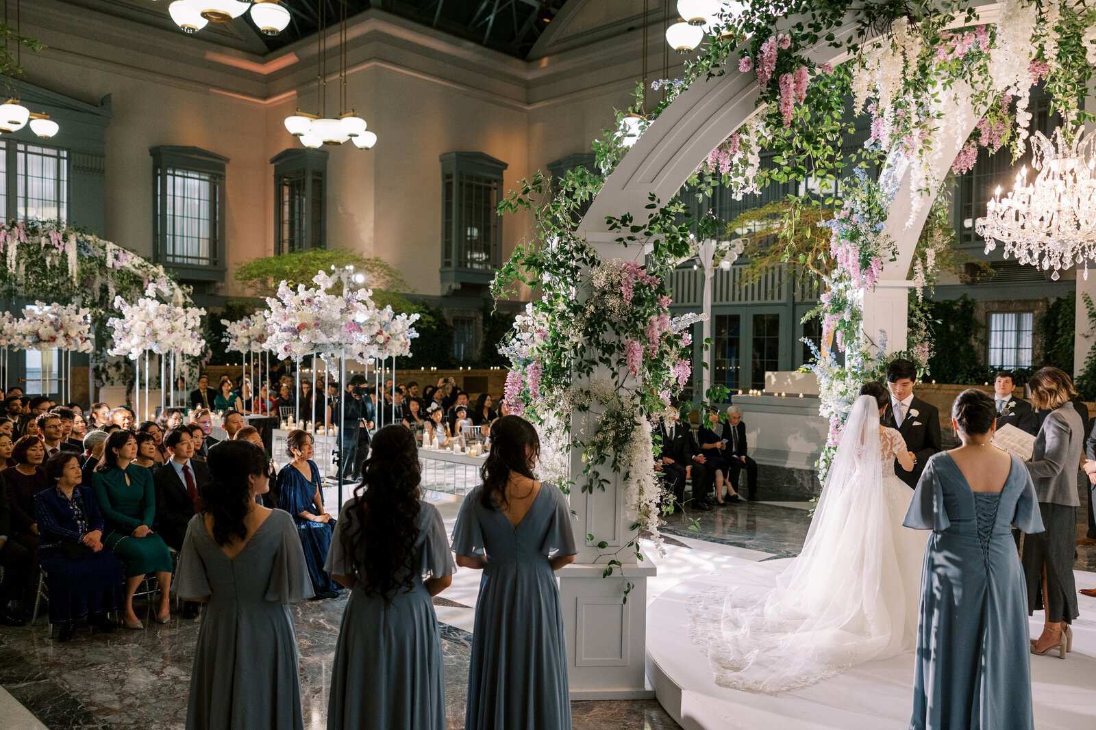 Colour photo of a couple at a flower-covered arch with bridesmaids and seated guests watching.